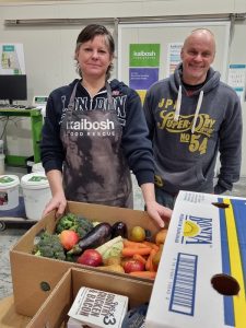 Two people stand with a large box of fresh produce, person at left wears an apron with Kaibosh Food Rescue on front.