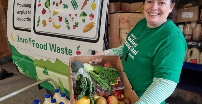 Woman holding box of produce stands in front of a Kaibosh food rescue van next to crates of milk bottles.