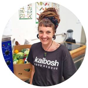 Woman smiling, wearing a Kaibosh t-shirt stands in front of a box of vegetables on a bench.